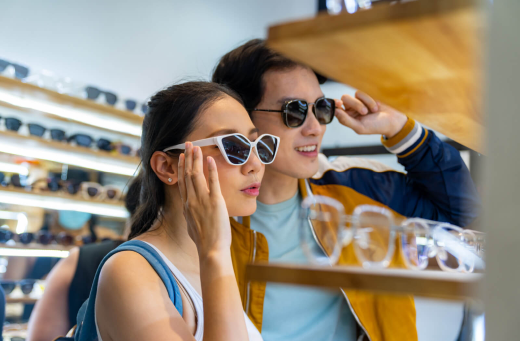 A young couple each trying on a pair of sunglasses in a store and looking in the mirror.