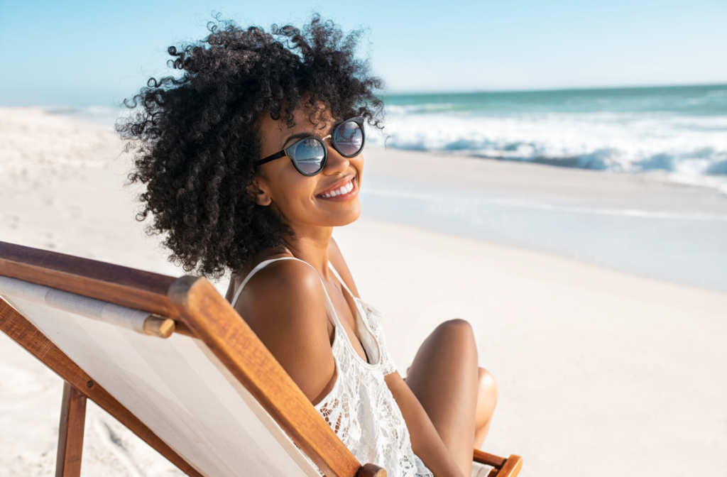 A young woman sitting on a lounge chair on the beach wearing sunglasses and smiling.
