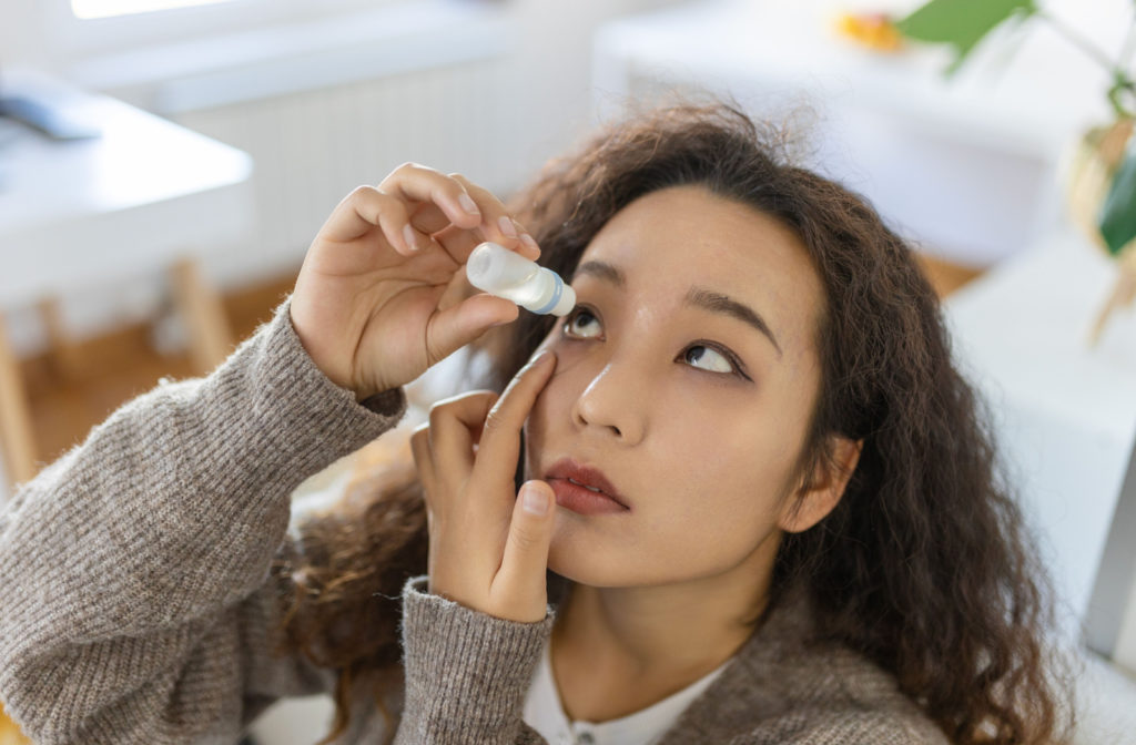 A curly-haired young woman applying eye drops to her right eye.