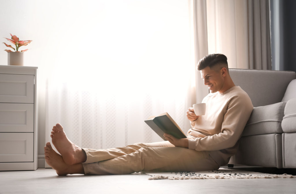A man reading a book while sitting on the living room floor and enjoying a cup of a hot beverage.