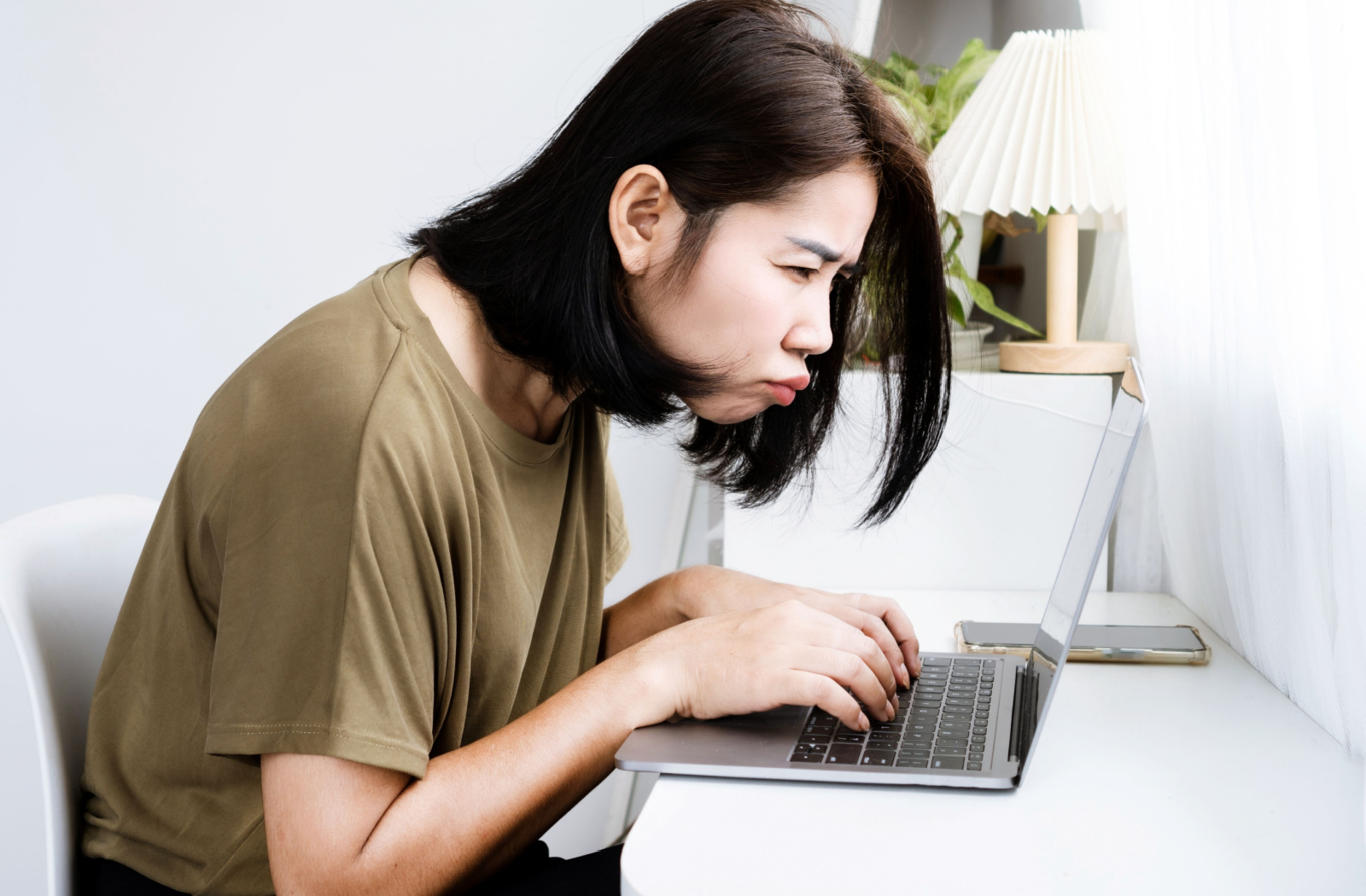 A woman hunched over while working on a laptop, leaning in and squinting to see the screen.