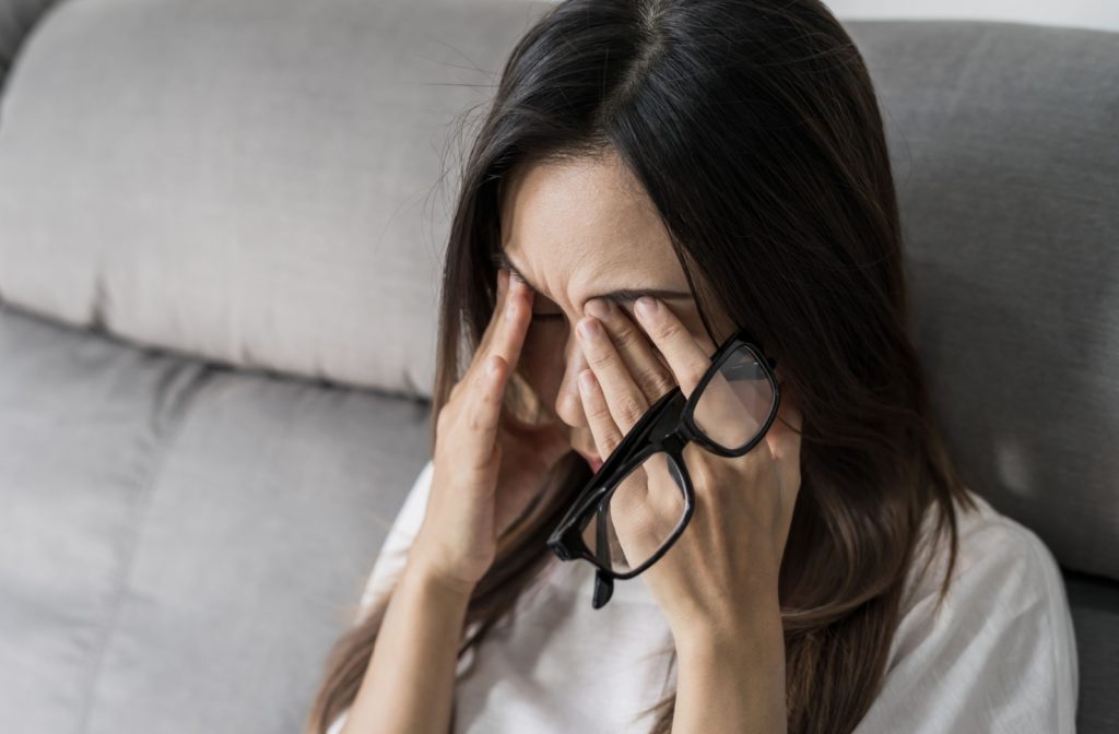 A person sitting on a couch has taken off their glasses to rub their dry and irritated eyes.