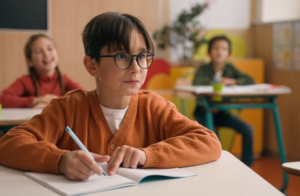 A young child wearing glasses and an sweater sits at a classroom desk, writing in a notebook.
