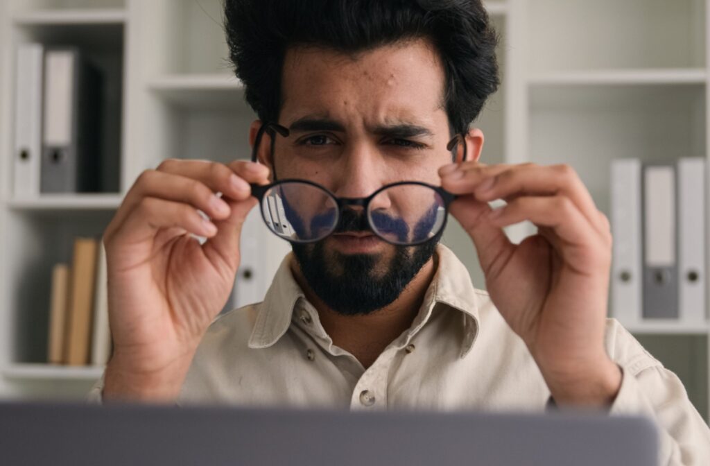 A person with a beard lifts his glasses while looking at a laptop screen, appearing to squint or concentrate.