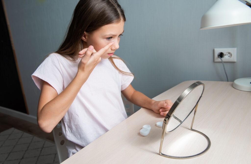Preteen age girl putting on contact lenses in front of a desk mirror.