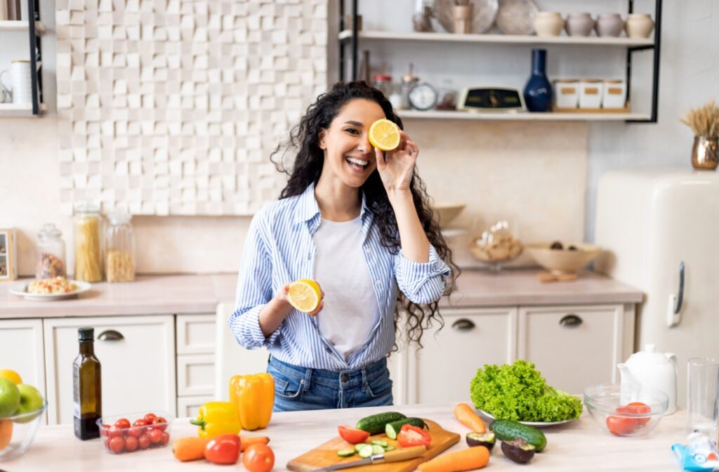 A person in the kitchen cooking with eye healthy fruits and vegetables, holding up an orange over their eye.