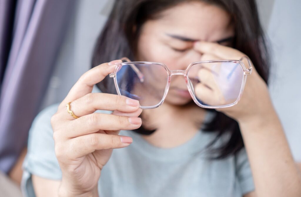 A person holding out their glasses in one hand and rubbing their eyes with the other.