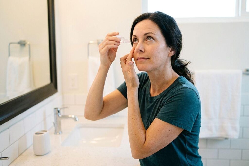 Adult applying eye drops to their eye while standing at a bathroom sink, holding a small dropper bottle, looking upward.